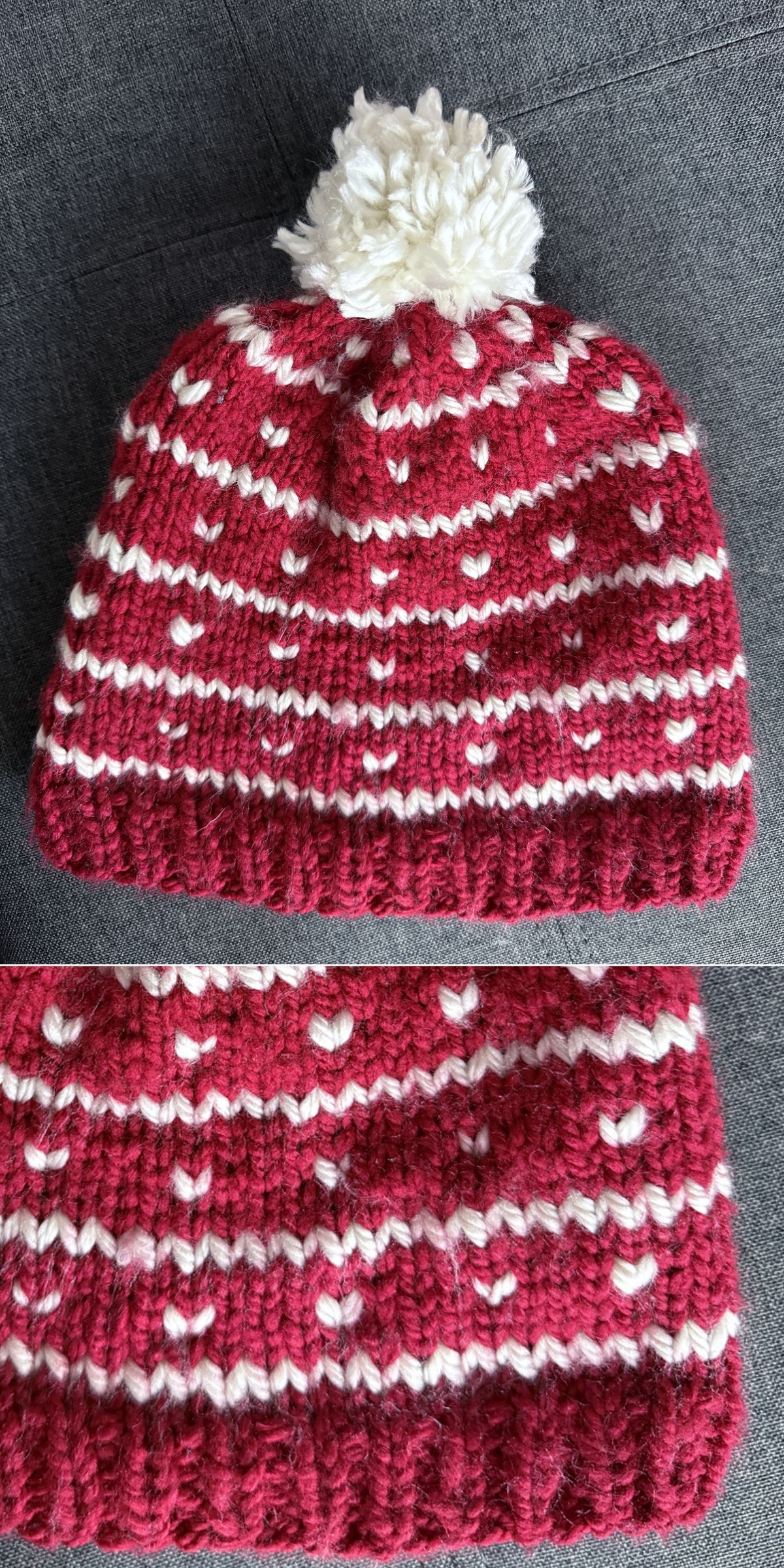 A knitted red beanie with white patterns, including a heart motif, and a white pom-pom on top, displayed on a gray fabric surface.
