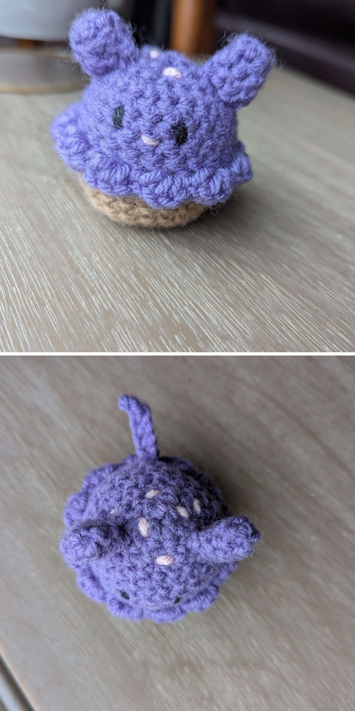A small crochet cat amigurumi plush figure with purple yarn, black eyes, two ears, and a tail, sitting on a wooden surface.