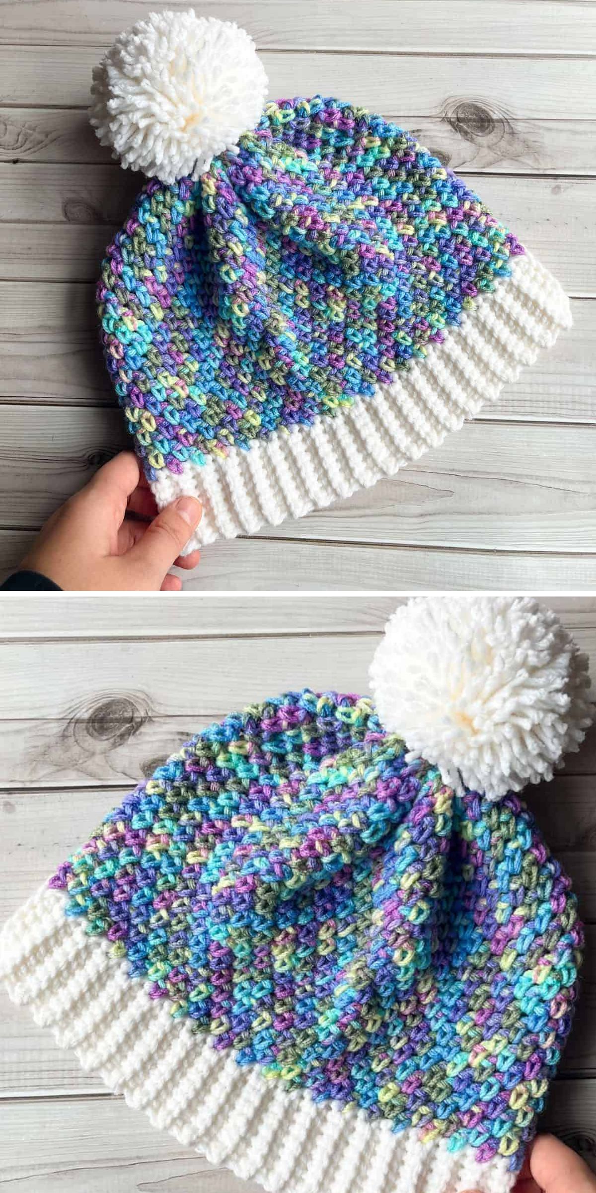 A hand holds a cozy crochet beanie with a white ribbed brim, a multicolored body in blue, green, and purple, and a large white pom-pom on top, displayed on a wooden surface.