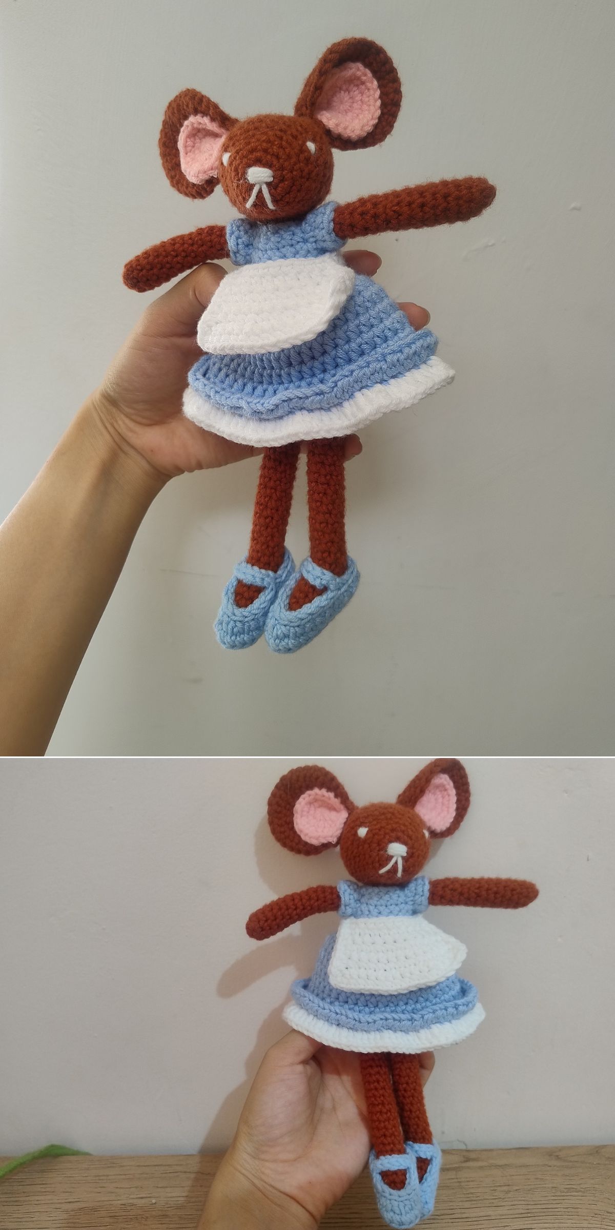 A hand holds an adorable amigurumi mice doll, crocheted in brown with pink ears, a blue dress, white apron, and matching blue shoes against a plain background.