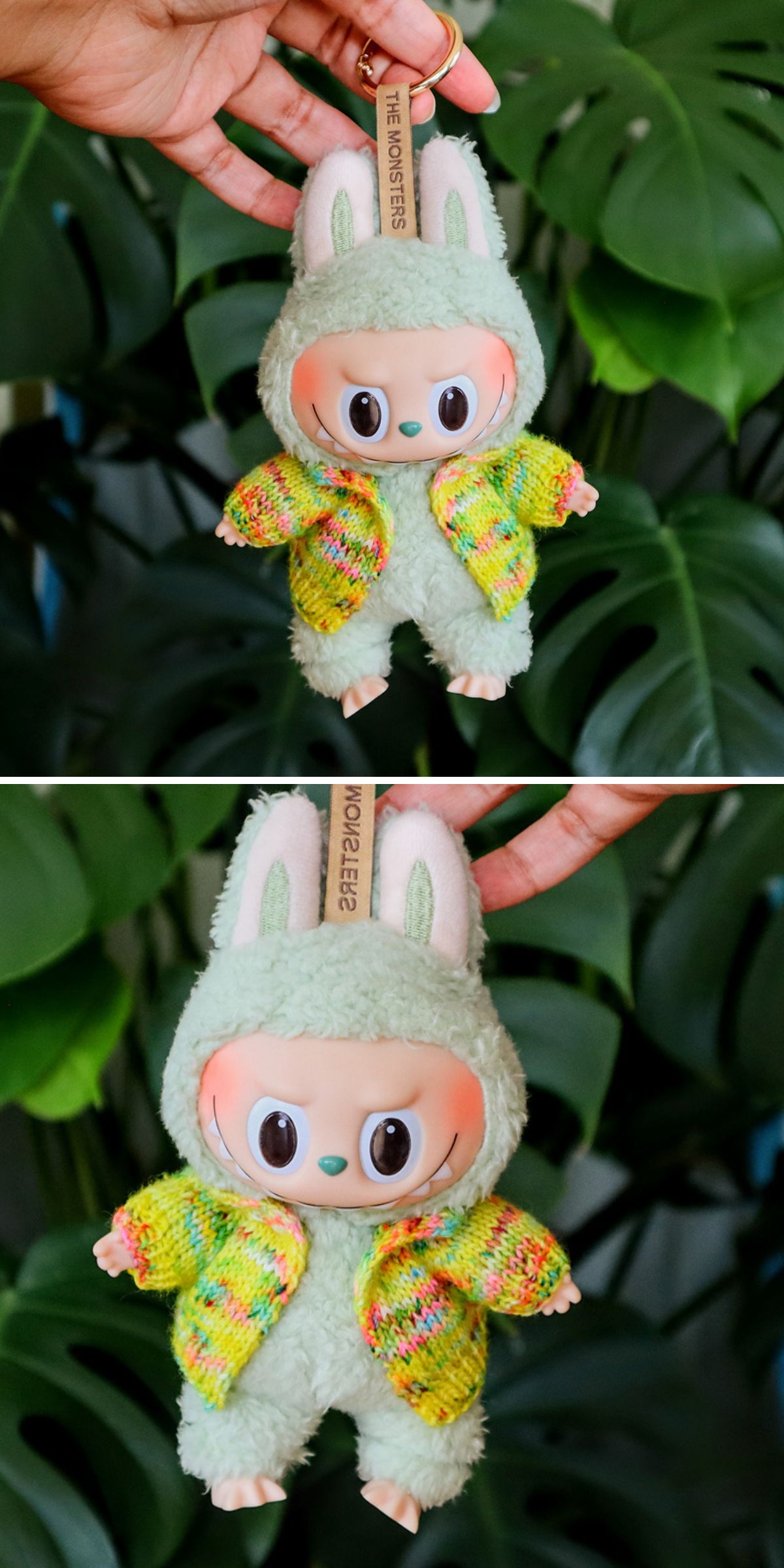 A hand holds a plush Labubu toy with a bunny hood, big eyes, and a colorful knitted cardigan — perfect for those searching for adorable knitted clothes for dolls — set against vibrant green leafy plants.