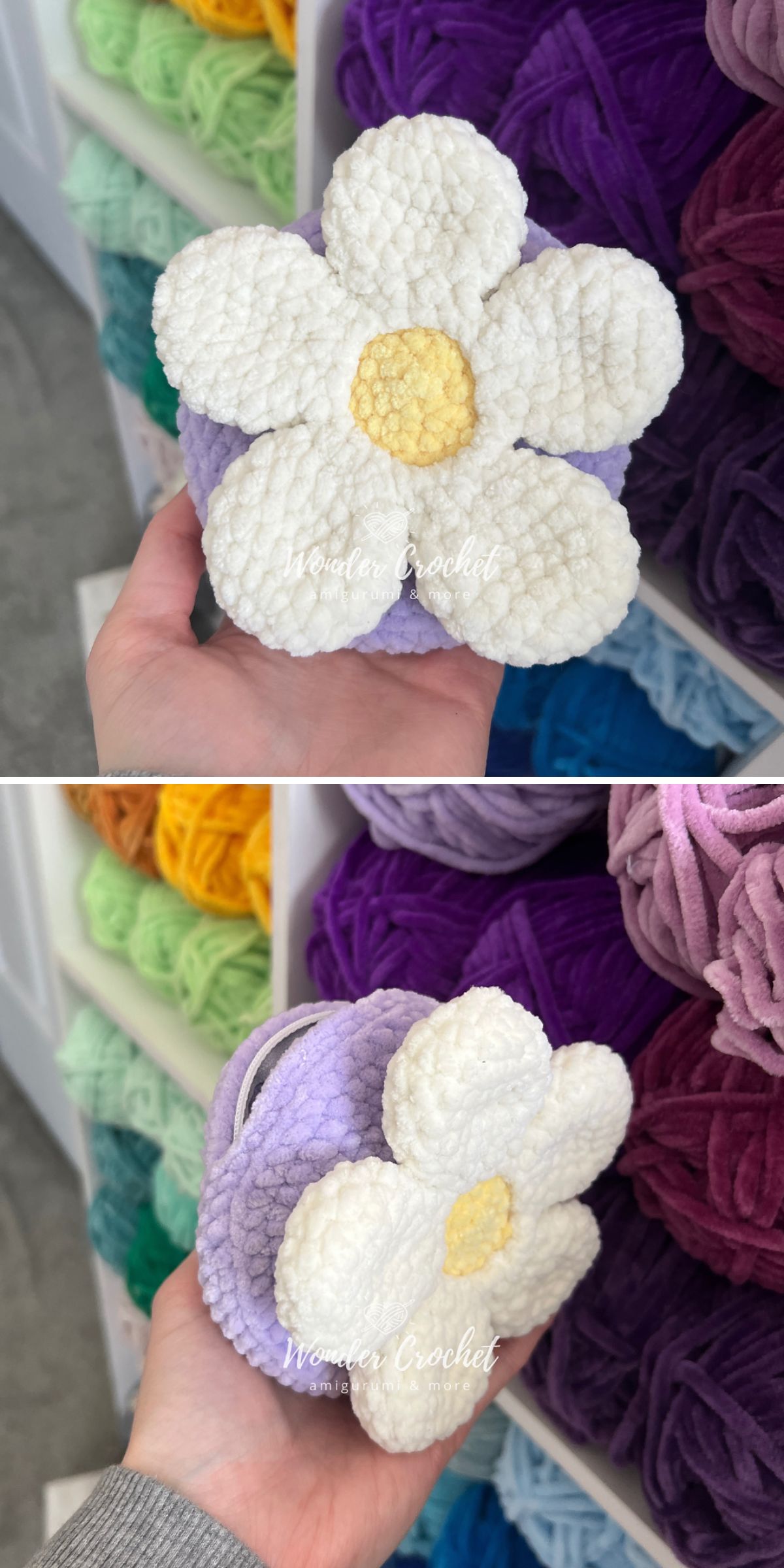 A hand holds a crocheted white and yellow flower with a purple base, possibly part of a crochet pouch, in front of shelves filled with colorful yarn balls.