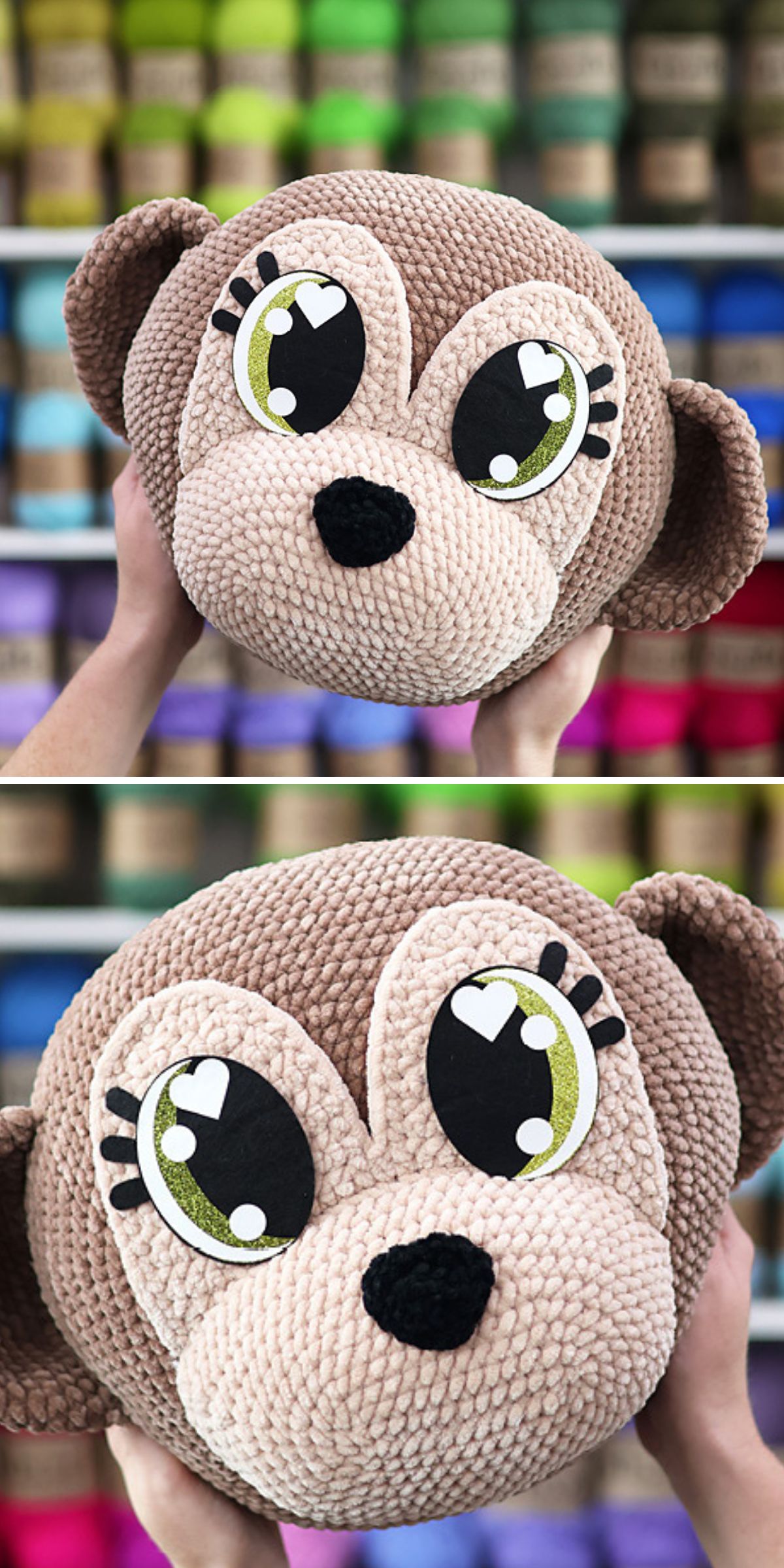 Two hands hold a crochet monkey plush toy shaped like a monkey's head in front of shelves of colorful yarn spools. The monkey has large green eyes with heart-shaped highlights.