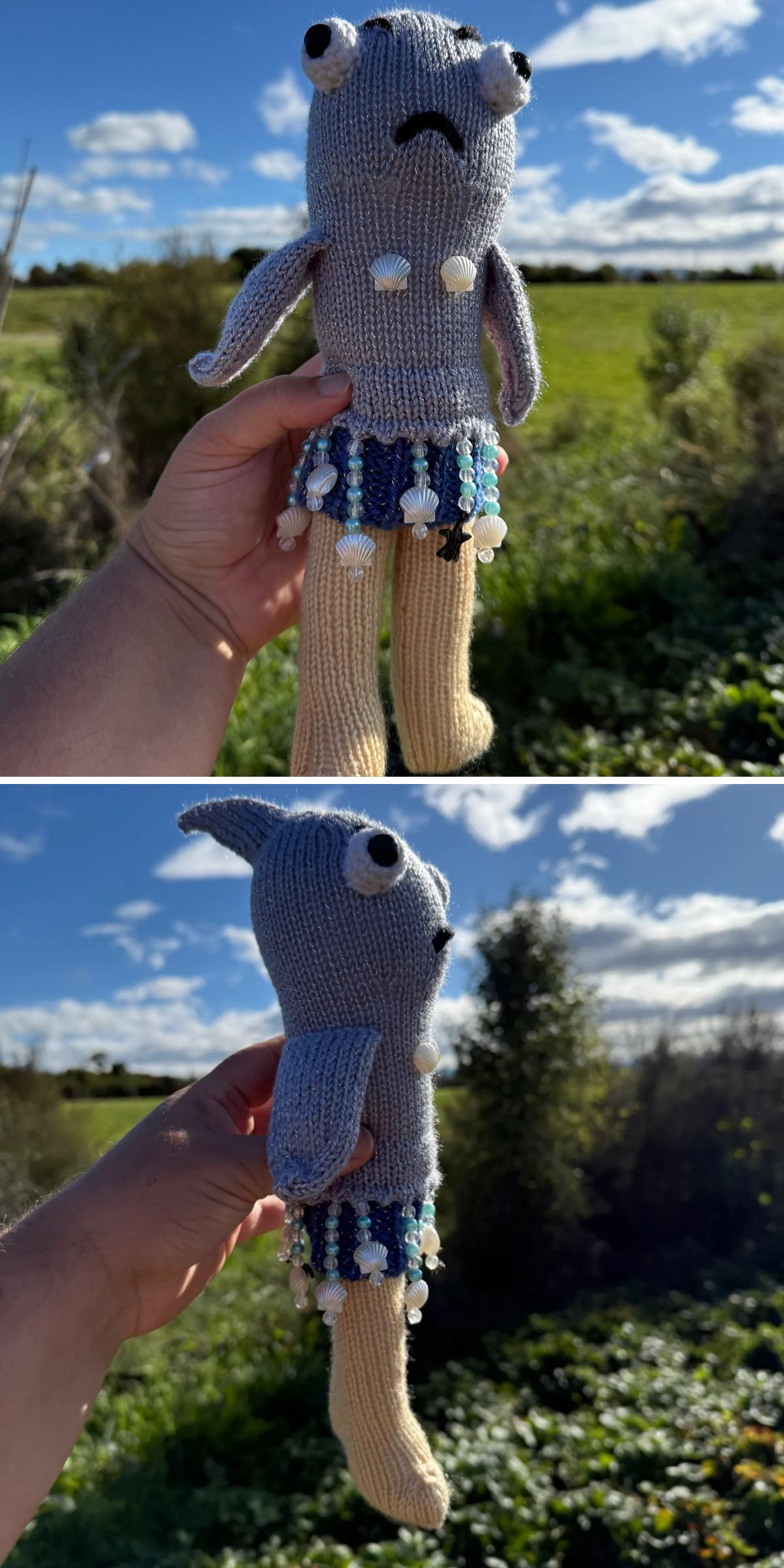 A hand holds a charming knitted toy doll with beige legs, a gray body, large eyes, and a blue skirt adorned with white beads. The background features greenery and a blue sky with clouds.