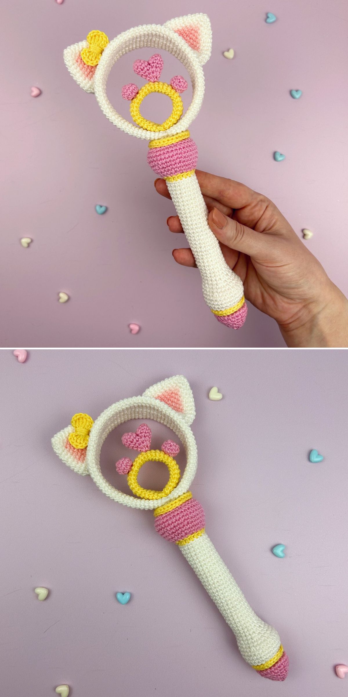 A hand holds a crochet baby toy — a cat-themed rattle with pink, yellow, and white details, cat ears, and a paw print. Small heart-shaped beads are scattered on the pink background.
