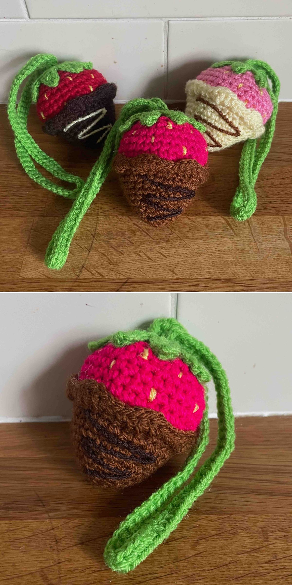 Three crochet strawberries with green stems, designed to look like chocolate-dipped treats, are displayed on a wooden surface against a white tiled background.