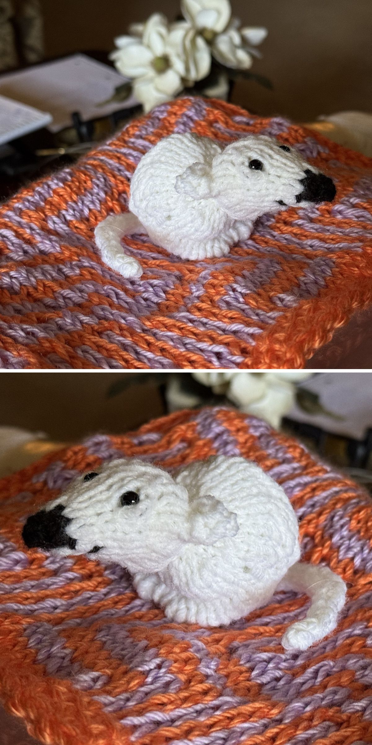 A small, white, whimsical knit toy with a black nose rests on a purple and orange knitted blanket; delicate white flowers bloom in the background.