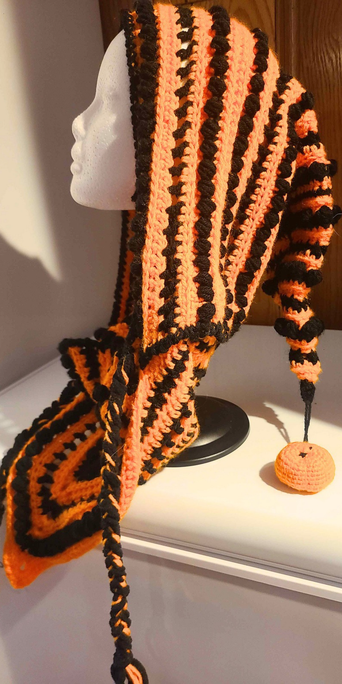 A crocheted Halloween hat styled as a striped orange and black hood on a mannequin head, featuring a long tail tipped with a small crocheted pumpkin.